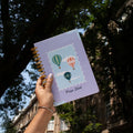 Hand holding hot air balloon notebook with name 'Priya Shah'